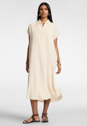 Kvinde iført en løs, kortærmet, elfenbensfarvet midi-kjole med krave, kombineret med mørkebrune sandaler og guldbøjler på det ene håndled.
