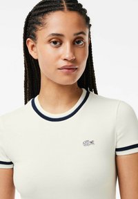 Camisa de manga corta color crema con cuello y puños de canalé en navy; presenta un pequeño logo bordado de cocodrilo en el lado izquierdo del pecho.