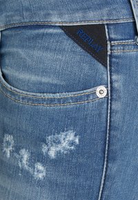 Jeans de mezclilla azul claro con textura desgastada, bolsillo con tela negra en contraste y logo azul "REPLAY", y herrajes plateados.