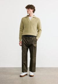 Νεαρός άνδρας με σγουρά μαλλιά, φορώντας μπεζ πουλόβερ με υφή, λευκό εσώρουχο, σκούρα καφέ παντελόνια, μαύρη ζώνη και λευκά αθλητικά παπούτσια, στέκεται σε γκρι πάτωμα.