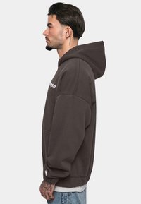 Dark brown hoodie with a spacious front pocket, ribbed cuffs, and a large hood, featuring white logo text on the front. Casual fit.