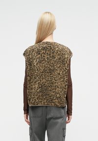 Γιλέκο από συνθετική γούνα σε καφέ χρώμα με υφή λεοπάρ μοτίβου, με στρογγυλή λαιμόκοψη και χωρίς μανίκια, φορεμένο πάνω από καφέ μπλούζα με μακρύ μανίκι.