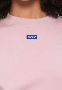 Camiseta de manga corta color rosa con textura acanalada y escote redondo. Presenta un logo rectangular azul con la palabra "HUGO" en el pecho.