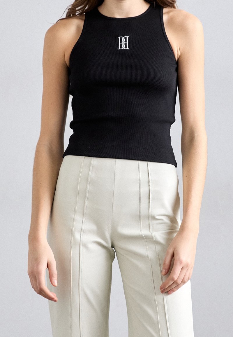 Femme portant un top noir ajusté sans manches avec un logo blanc et un pantalon beige taille haute avec des détails de coutures verticales.