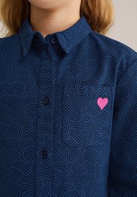 Camicia blu navy con un motivo geometrico a forma di cuore di colore blu chiaro, con un cuore ricamato di colore rosa sulla tasca del petto e chiusura con bottoni.