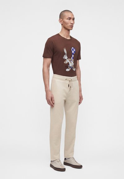 Άνδρας που φορά καφέ T-shirt με γραφικό Bugs Bunny, μπεζ παντελόνι με κορδόνι και μπεζ αθλητικά παπούτσια, στέκεται μπροστά σε απλό φωτεινό φόντο.