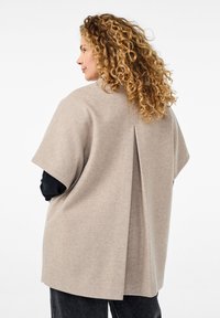 Top in misto lana beige con maniche corte, caratterizzato da una piega sul retro e una vestibilità morbida. Texture liscia, design minimale e colore neutro.