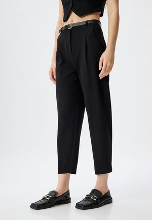 Black tailored trousers with pleats, cropped length, and a sleek belt. Paired with black loafers featuring textured finish and gold hardware.