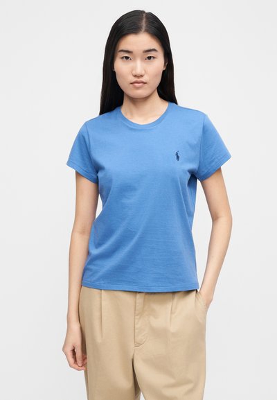 Kék pamut póló kerek nyakkivágással, rövid ujjú, bal oldali mellrészen kis sötét logóval; bézs nadrággal párosítva.