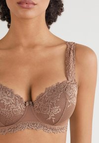 Koronkowy stanik w miękkim brązie z misternymi wzorami kwiatowymi, gładką siateczkową podstawą i dekoracyjnymi ramiączkami z falbaniastymi krawędziami.
