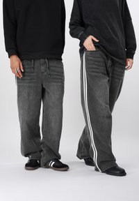 Μαύρο φούτερ με κουκούλα συνδυασμένο με γκρι τζιν παντελόνι φαρδιάς γραμμής με λευκές ρίγες στα πλαϊνά. Και οι δύο φορούν μαύρα παπούτσια· το ένα έχει μαύρη σόλα, το άλλο καφέ σόλα.