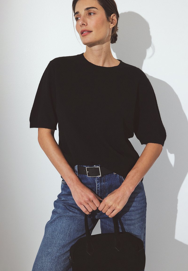 Femme portant un haut noir à manches courtes et un jean bleu, tenant un sac à main noir, se tenant contre un mur blanc avec une ombre douce.