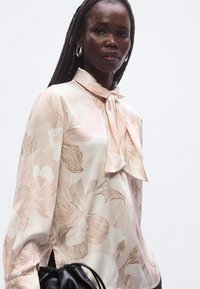 Halenka z lehké tkaniny s krémovým pozadím a velkými růžovými květinovými vzory, se zavazovacím límcem a dlouhými rukávy.