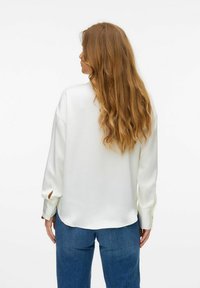 Witte blouse met lange mouwen, een gladde textuur en zijopeningen bij de manchetten, gecombineerd met blauwe jeans, gezien van achteren.