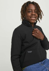 Svart zip-up jacka med hög krage, med en framficka där geografiska koordinater är tryckta. Jackan har en slät textur och en minimalistisk design.