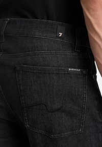 Zwarte denim jeans met een strak, gladde textuur. Opvallende details zijn een zak met een golvende stiknaad en een merketiket op de tailleband.