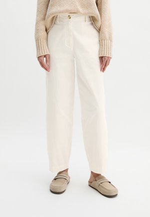 Osoba ubrana w szerokie, kremowe spodnie, beżowy dziergany sweter oraz beżowe mokasyny na płaskim obcasie, stojąca na jednolitym białym tle.