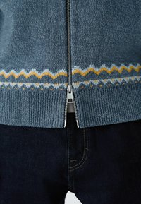 Kék kötött, zipzáros pulóver bordás szegéllyel és a alján kontrasztos sárga és fehér cikk-cakk mintával. Sötét farmernadrággal párosítva.
