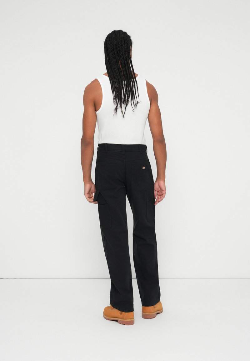 Pantalons cargo noirs avec des poches latérales, fabriqués en tissu durable. Associés à un débardeur blanc et des bottes de travail marron. Fond uni.