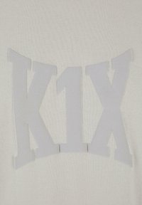 Balts džemperis ar teksturētu, izvirzītu "K1X" logotipu nedaudz gaišākā pelēkā krāsā, centrēts uz gludas adītas auduma.
