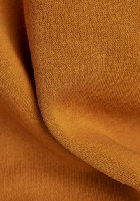 Tejido de algodón en un rico color mostaza, con una textura de punto fino y suaves pliegues, que muestra sutiles variaciones en el tono.