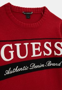 Punane kudum särk ribatud ümarakaelusega, millel on suured valged "GUESS" tähed ja "Authentic Denim Brand" kiri musta riba üle.