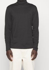 Maglione a collo alto nero con maniche lunghe in un tessuto liscio e elasticizzato, dal taglio slim, con un logo discreto sul petto. Pantaloni di colore chiaro.