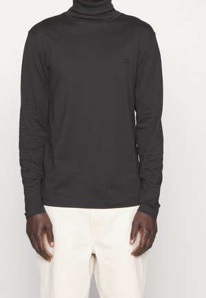 Schwarzes Rollkragenlangarmshirt aus einem glatten, dehnbaren Stoff mit schmaler Passform, das ein dezentes Logo auf der Brust trägt. Helle Hose.