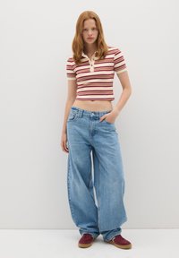 Joven mujer de pie, vistiendo una camiseta polo de rayas corta, jeans de pierna ancha de color azul claro y zapatillas color burdeos, frente a una pared lisa.