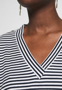 Tessuto a righe in bianco e nero con scollo a V, caratterizzato da un knit testurizzato e un dettaglio di colletto a coste. Orecchino di colore dorato visibile.