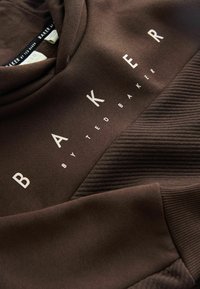 Hnedá mikina s kapucňou, ktorá obsahuje textúrovanúplet, biele logo "BAKER by TED BAKER" a mäkkú látku. Detaily tvoria rebrované manžety a lem.
