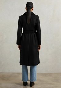 Naine seisab seljaga, kandes musta vöömehega kasukat, helesiniseid teksaseid ja musti kontsad, lihtsa beeža tausta ees.