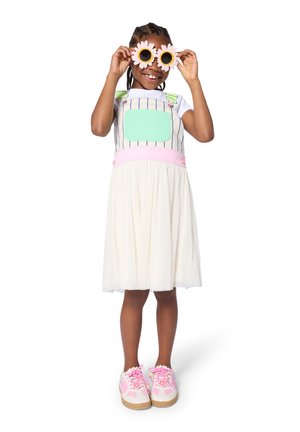 Fille souriante tenant des lunettes de soleil en forme de fleur rose près de son visage, portant une robe blanche avec une salopette pastel et des baskets roses.