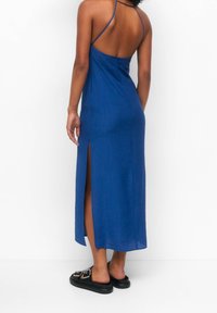 Femme portant une robe bleu sans manches avec des bretelles dans le dos et une fente latérale, associée à des sandales noires à enfiler, debout sur un fond blanc.