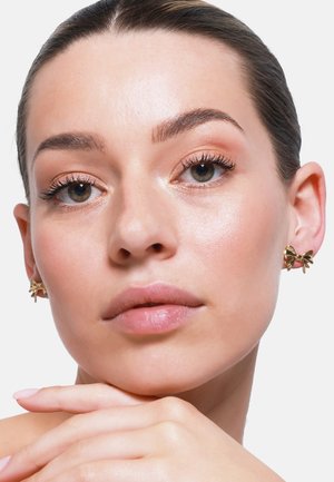 Gouden strikvormige oorbellen sieren de oren van het model. Ze heeft een gladde teint, met zachte verlichting die haar gelaatskenmerken en sieraden accentueert.