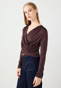 Bruine wrap-style lange mouw top met een gedrapeerde voorkant en aansluitende mouwen, gecombineerd met hoog-getailleerde donkerdenim jeans met zakken.