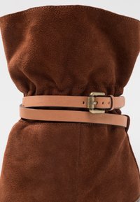 Brun mocka knähöga stövlar med samlat topp, utrustade med ett bälte i läder i tanfärg och en brass spänne detalj. Slät textur och enkel design.