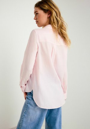 Neiu laineliste blondide juustega, kes kannab heleroosat pikkade varrukatega särki ja lahtisi siniseid teksapükse, poseerib külili tavalise tausta ees.