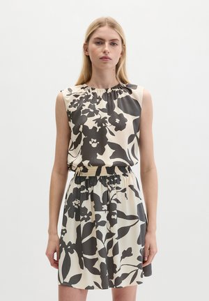 Junge Frau trägt ärmelloses beiges Kleid mit schwarzem Blumenmuster und elastischer Taille, steht vor schlichtem weißem Hintergrund.