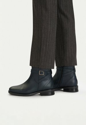 Dark blue leather ankle boots with side buckle and zipper worn with dark pinstriped trousers.