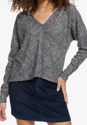 Kvinde iført en grå tekstureret V-hals sweater og en mørkeblå fløjlsnederdel, hånden hvilende på nederdelens talje.