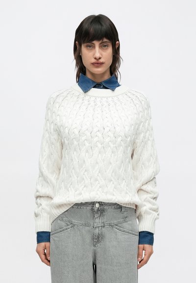 Pull crème en maille torsadée avec un col rond, associé à une chemise en denim bleu en dessous. Le modèle porte un jean gris clair à jambes larges.