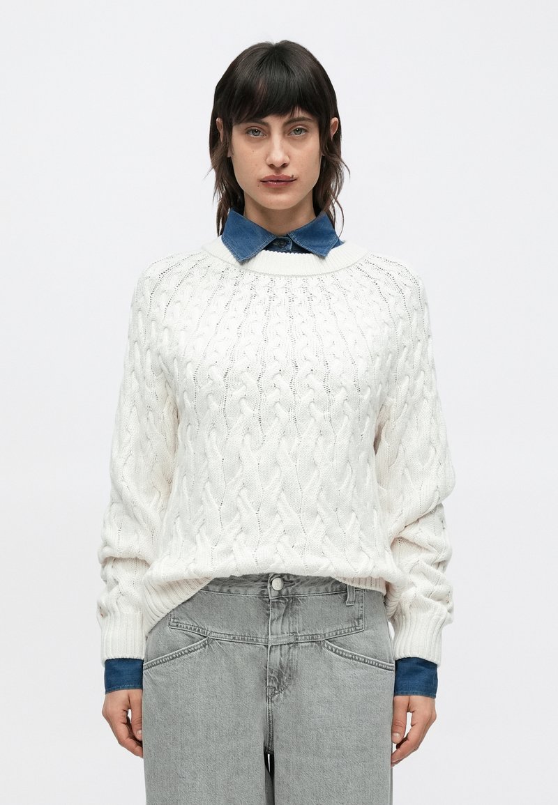 Κρεμ πουλόβερ με πλέξη καλωδίου και στρογγυλή λαιμόκοψη, συνδυασμένο με μπλε τζιν πουκάμισο από μέσα. Το μοντέλο φορά ανοιχτό γκρι ευρυκάβαλα τζιν παντελόνια.