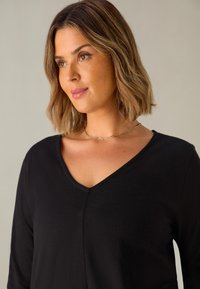 Černý top s výstřihem do V a dlouhými rukávy, vyrobený z měkké látky. Obsahuje jemný ševový detail na přední straně. Modelka nosí jednoduchý zlatý řetízek.