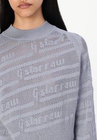 Pull en tricot gris clair avec un col haut, présentant un motif texturé de lignes horizontales et de lettres. Le tissage ouvert offre une respirabilité.