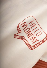 T-paita puuvillasta, jossa on vaalean kermaan väri ja punainen graafinen kuvio, jossa on puhekupla, jossa lukee "HELLO MONDAY". Pehmeä pinta ja rento malli.