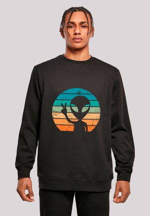 Junger Mann mit geflochtenen Haaren, der einen schwarzen Pullover mit buntem Retro-Alien-Design trägt und vor grauem Hintergrund ein Peace-Zeichen macht.
