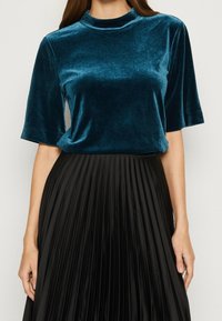 Sametový tyrkysový top s krátkými rukávy a vysokým výstřihem, doplněný skládanou černou sukní se lesklým povrchem a vertikálními pruhy.