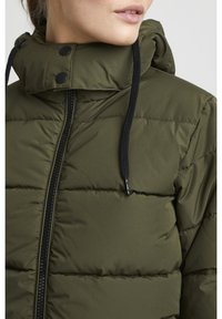 Grön vadderad jacka med hög krage, svart dragkedja, kontrasterande dragsnören och vadderad textur med horisontella sömmar.