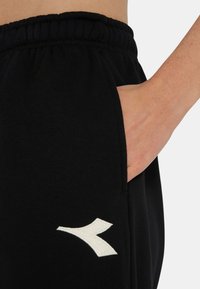 Pantaloni della tuta neri con vita elastica. Presentano una tasca laterale e un logo bianco ricamato sulla gamba sinistra. Tessuto dalla texture morbida.